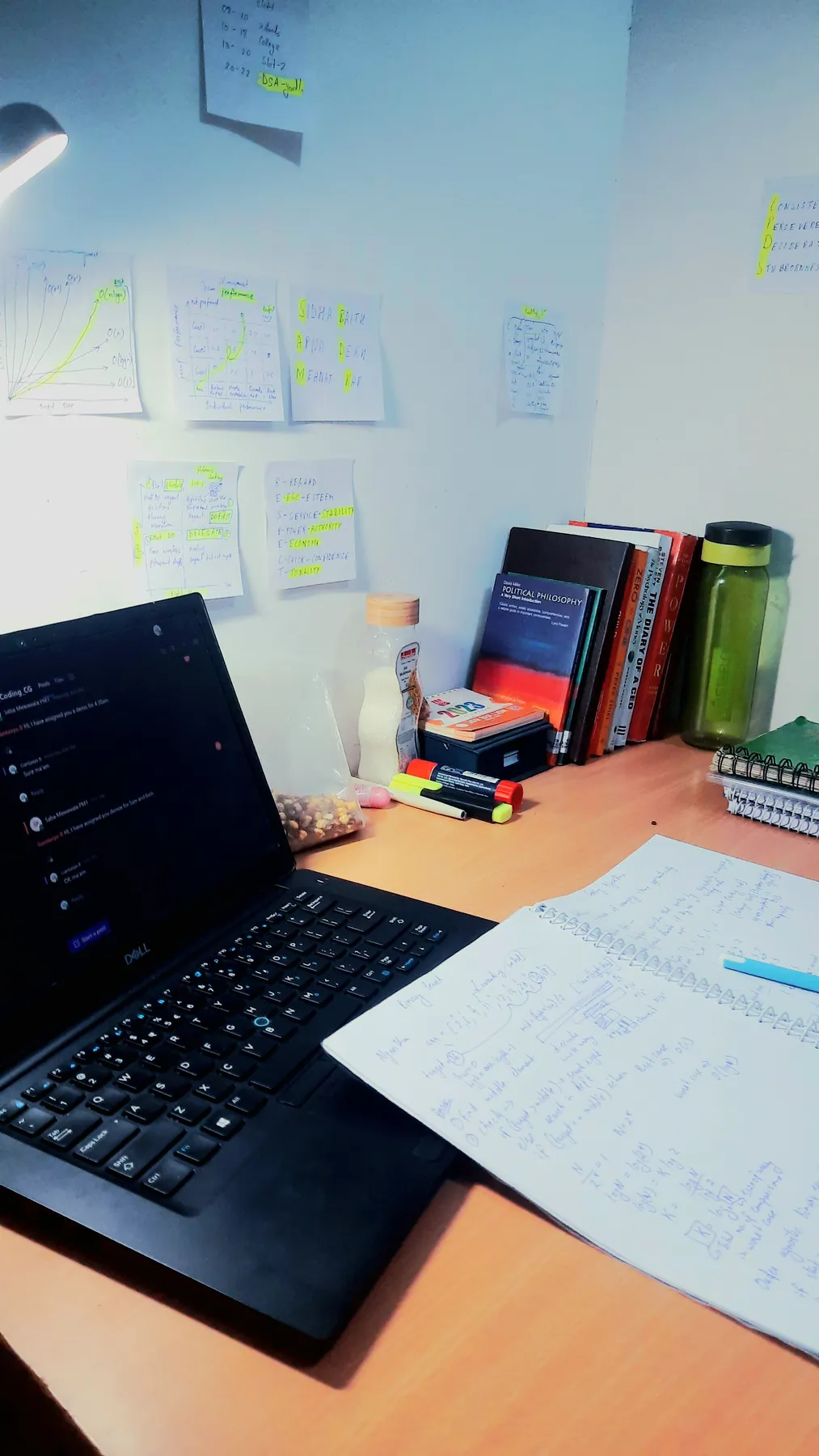 A desk at night: laptop, an open notebook of sketches, and sticky notes with diagrams on the wall